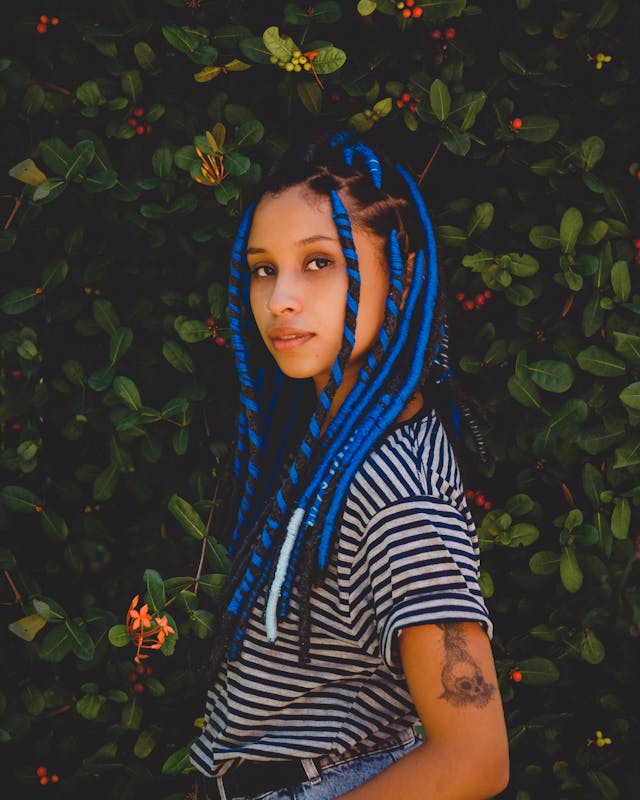 Young woman with long blue braids and striped shirt posing outdoors, showcasing professional women’s braiding services at Tru Hair Studio in Brighton, Colorado.